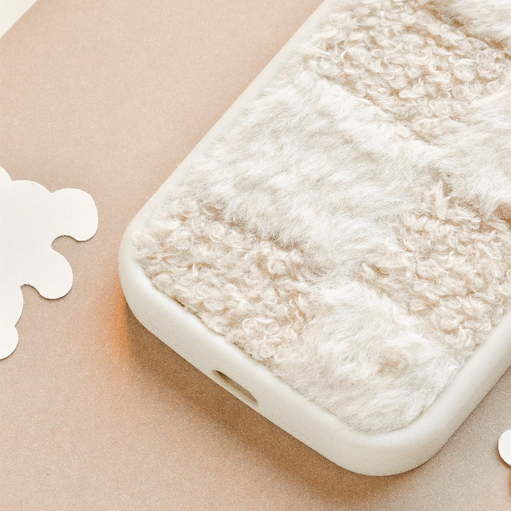 A close-up image of a Cosy Teddy Phone Case - Vanilla Biscuit from Coconut Lane, showcasing its soft, textured beige and white fur-like fabric covering. The case edges are smooth and white, providing a pleasing contrast to the cosy grip surface. The background features light, neutral tones and abstract shapes.