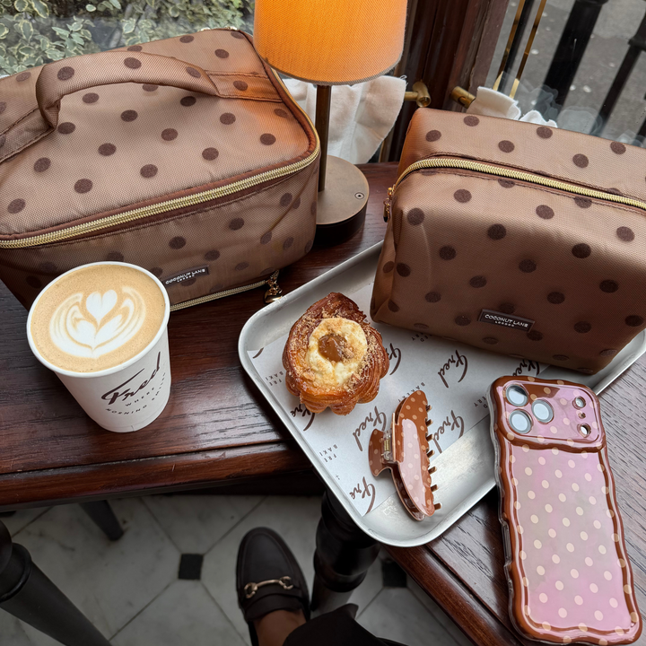 Flat lay photo in a cafe of the Dot Society Wavy Chocolate Brown collection. Including the Wavy chocolate brown case, with lighter brown polka dots, alongside the matching claw clip, makeup bag and vanity case. 