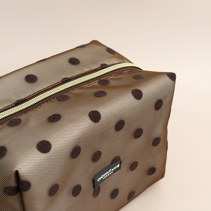 Top view of brown mesh makeup bag with dark brown polka dots, a gold top zip with a heart-shaped zip pull, and a black Coconut Lane London logo badge on the front, shown on a beige background.
