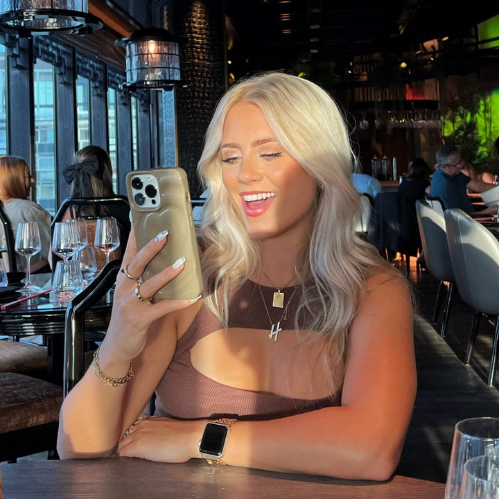 A woman with long blonde hair is sitting at a restaurant table, smiling at her smartphone adorned with Coconut Lane’s Melting Heart Phone Case in Biscuit Beige. She is wearing a brown sleeveless top, a gold necklace, and an extra protective smartwatch. The restaurant’s elegant decor and soft lighting create a warm ambiance, complete with wine glasses on the table.