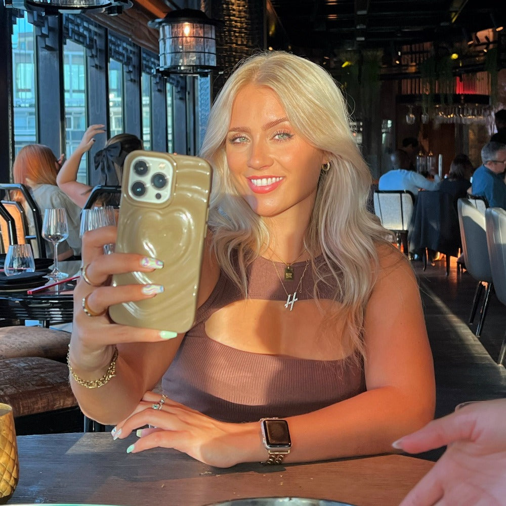 A woman with long blonde hair is taking a selfie in an elegant restaurant with large windows and ambient lighting. She is smiling and wearing a brown sleeveless top, an Apple Watch, and holding her phone adorned with the Melting Heart Phone Case in Biscuit Beige by Coconut Lane. Glasses and plates are on the table in front of her.