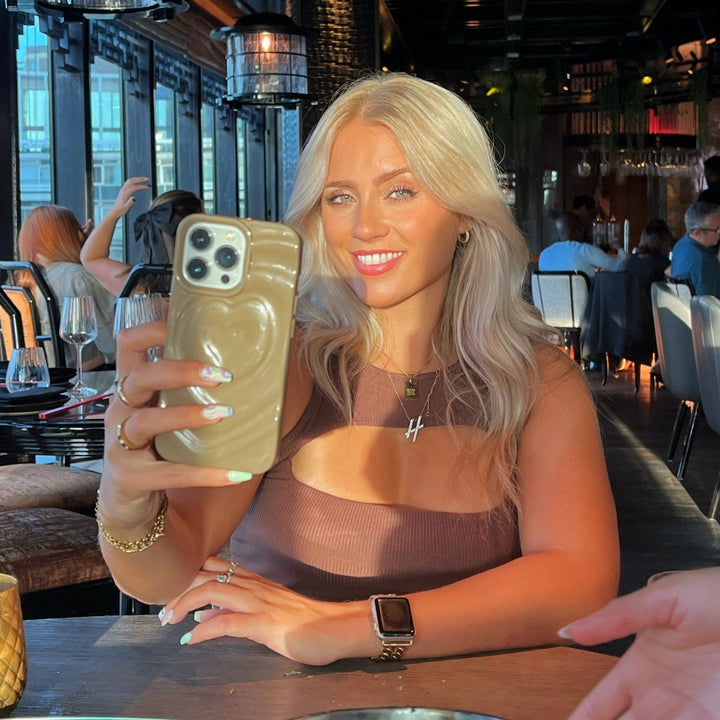 A woman with long blonde hair is taking a selfie in an elegant restaurant with large windows and ambient lighting. She is smiling and wearing a brown sleeveless top, an Apple Watch, and holding her phone adorned with the Melting Heart Phone Case in Biscuit Beige by Coconut Lane. Glasses and plates are on the table in front of her.