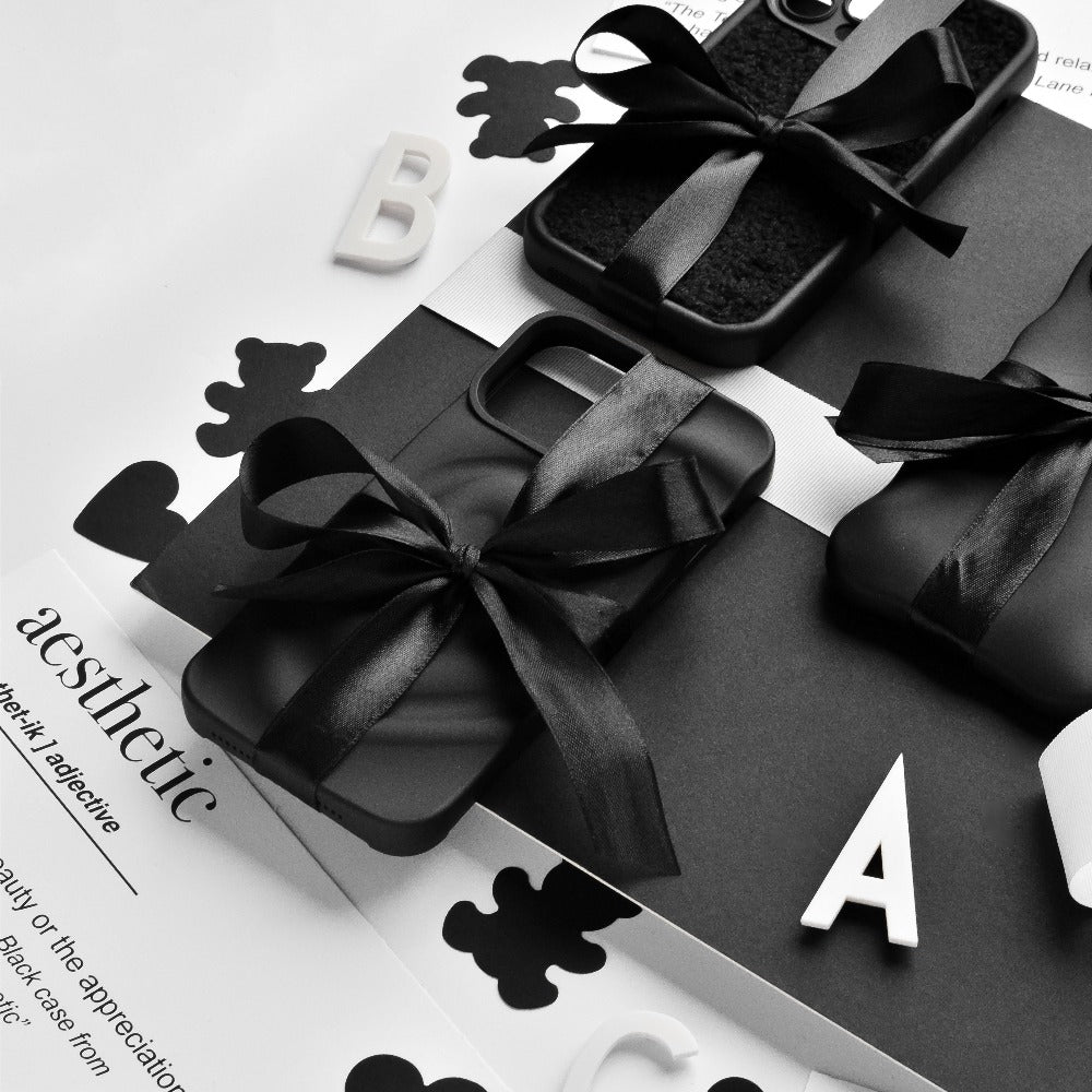 A monochrome photo featuring Coconut Lane's Melting Heart Phone Cases in Matte Black, elegantly tied with black ribbons. They are arranged on a black and white surface adorned with scattered letters and cut-out shapes, with the word "aesthetic" partially visible on a sheet of paper in the background.