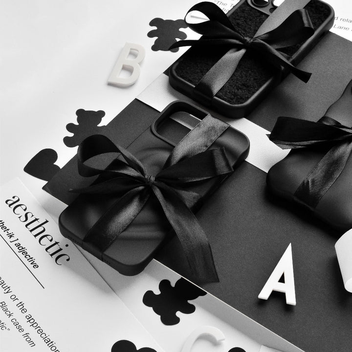 A monochrome photo featuring Coconut Lane's Melting Heart Phone Cases in Matte Black, elegantly tied with black ribbons. They are arranged on a black and white surface adorned with scattered letters and cut-out shapes, with the word "aesthetic" partially visible on a sheet of paper in the background.