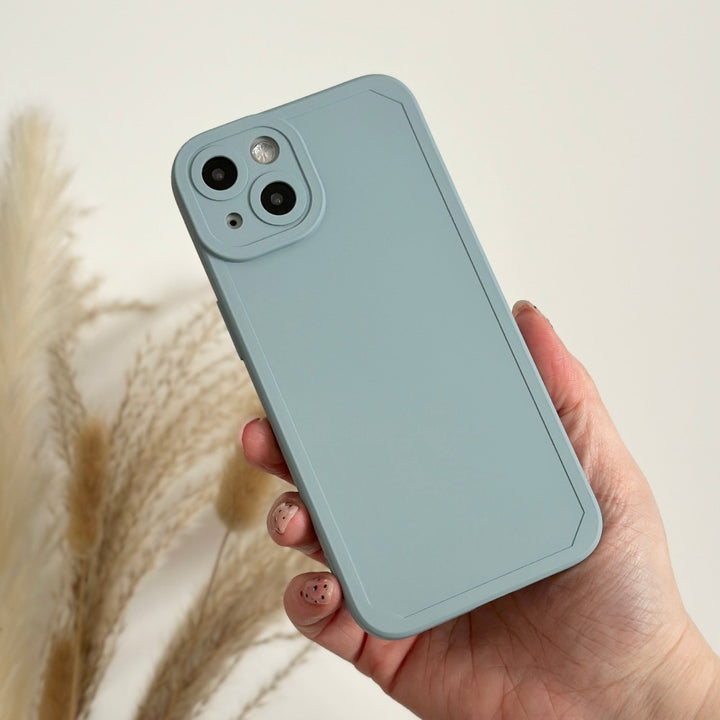 A hand is holding the NAKD Phone Case - Baby Blue by Coconut Lane, showcasing its minimalist design. Made of soft, flexible TPU, it fits snugly on the back of a phone and offers full camera protection for its dual rear cameras and flash. In the background, softly focused beige pampas grass stalks gently sway.