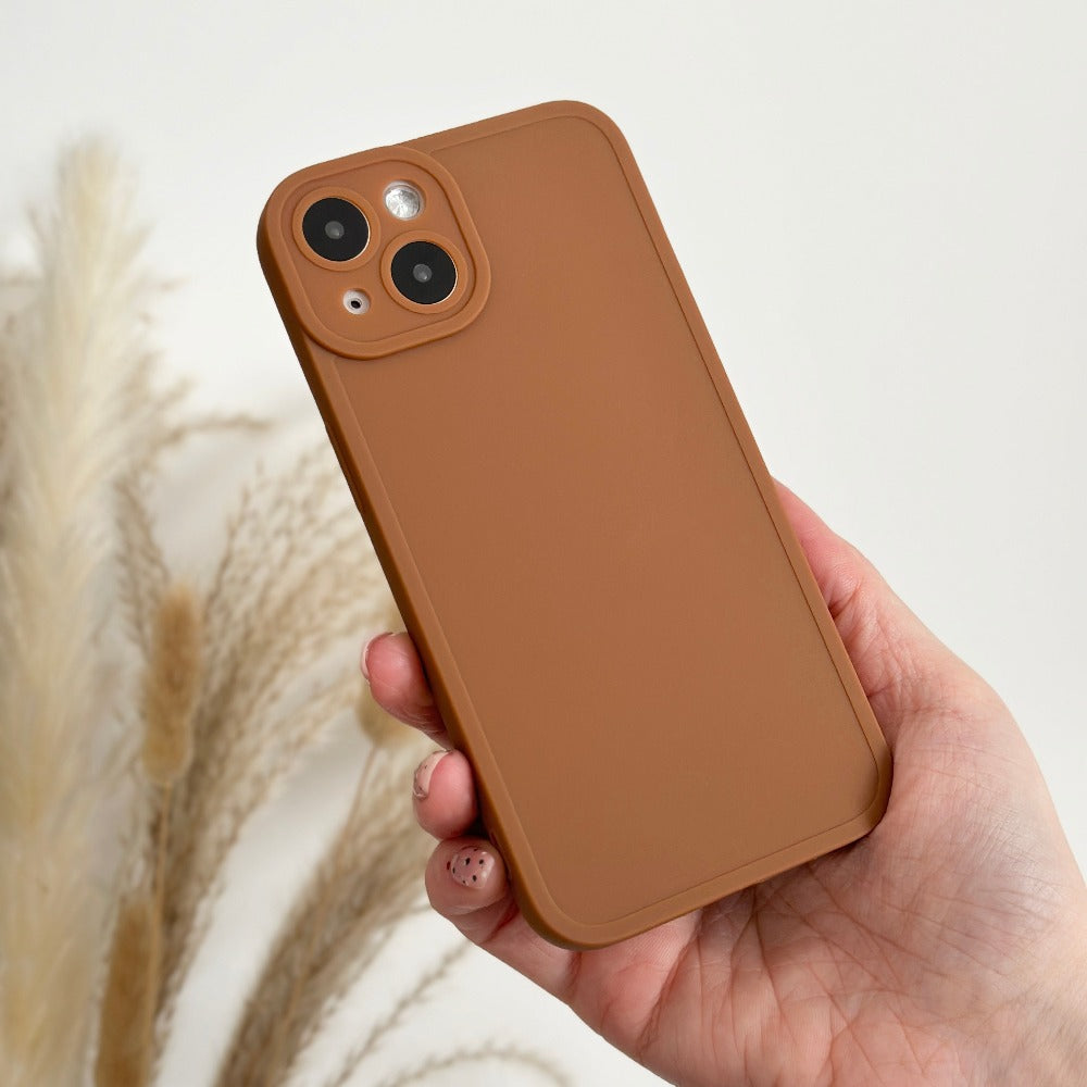 A hand holding a smartphone with a Coconut Lane NAKD Phone Case in Chocolate Brown. The phone features dual rear cameras and a minimalist design. In the background, decorative dried pampas grass stems stand against a light-colored wall.