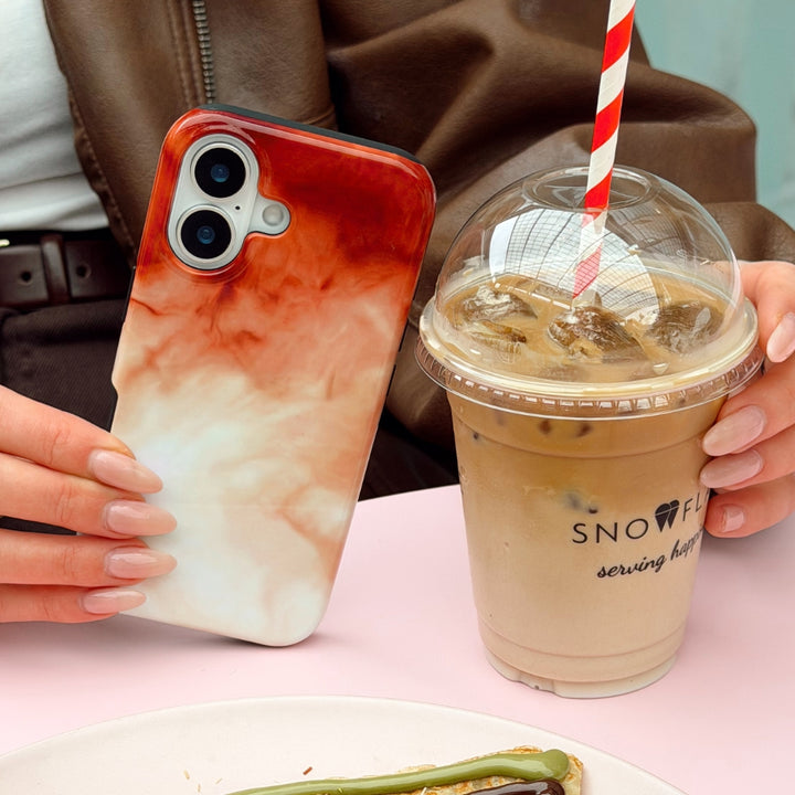 Phone case with a marble-style design held by a hand next to an iced coffee drink.