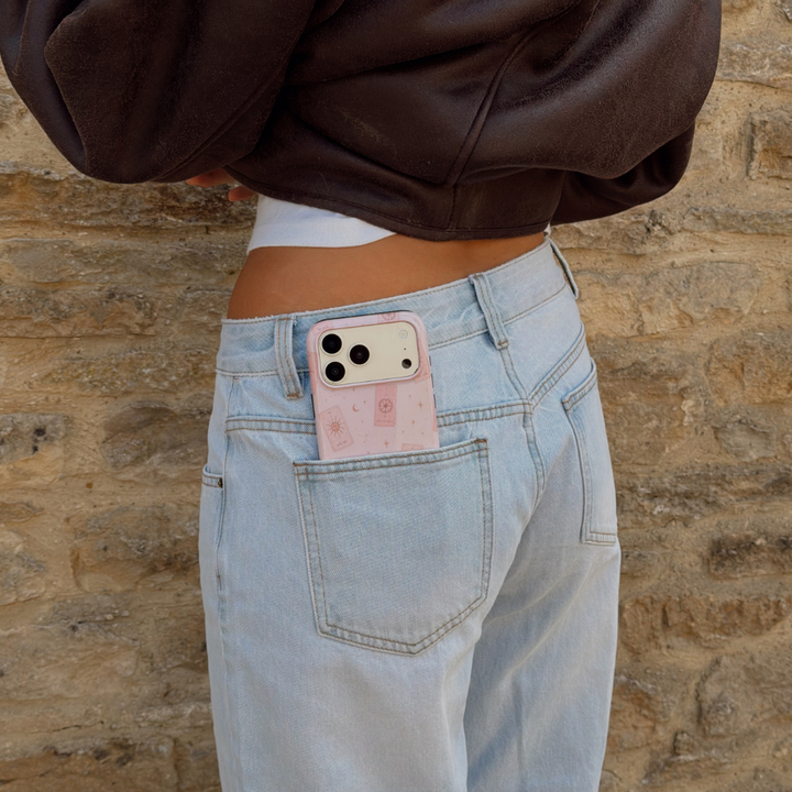 Woman wearing light blue jeans, showing a phone peaking out her back pocket, revealing a light pale pink phone case with tarot cards and tiny stars. The woman is wearing a brown aviator jacket. 