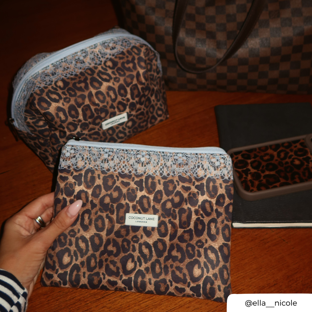 Woman holding the leopard print zip pouch with a pale blue lace trim along the top, a pale blue zip with a silver pull, and a white Coconut Lane London logo badge on the front. In the background is the matching makeup bag, alongside the mocha leopard phone case. 