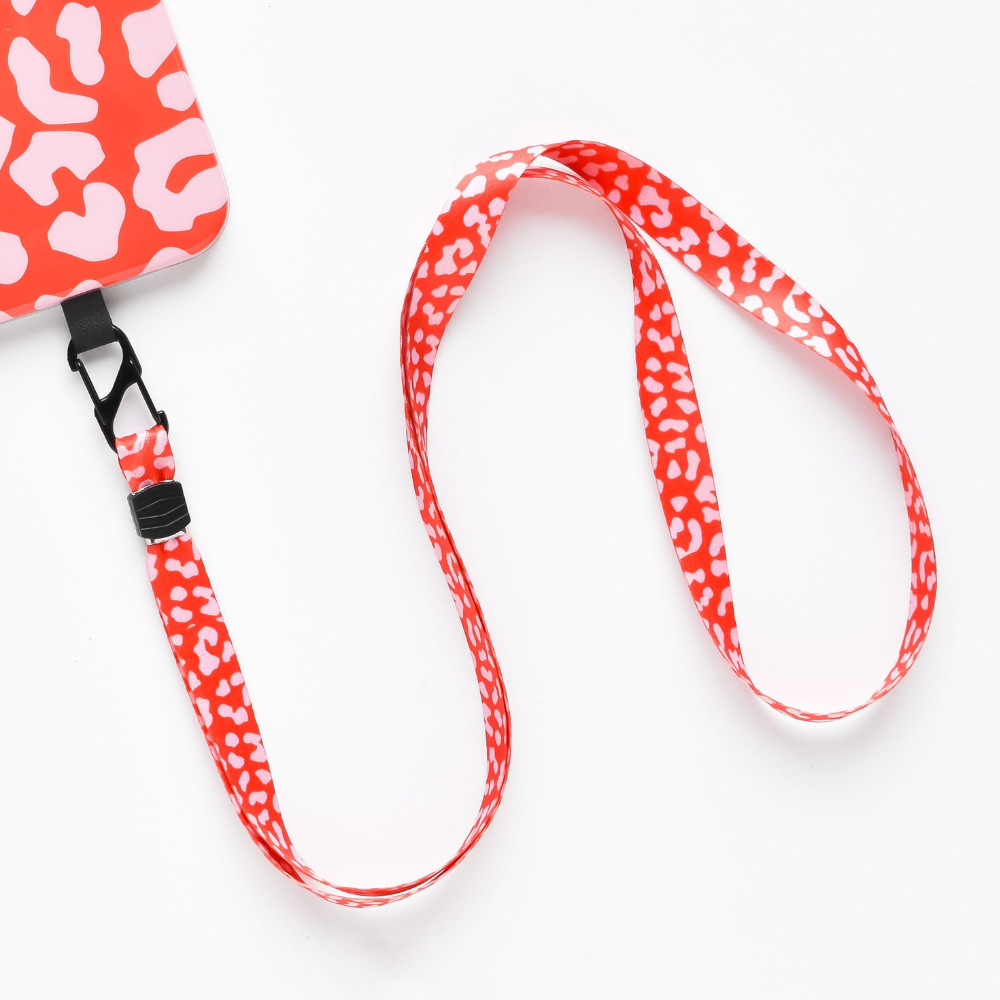A red Coconut Lane lanyard with a white leopard print pattern is attached to a matching Universal Phone Lanyard Patch Bundle in red and white. The lanyard features a black clasp connecting it to the phone security accessory. The background is a plain white surface.
