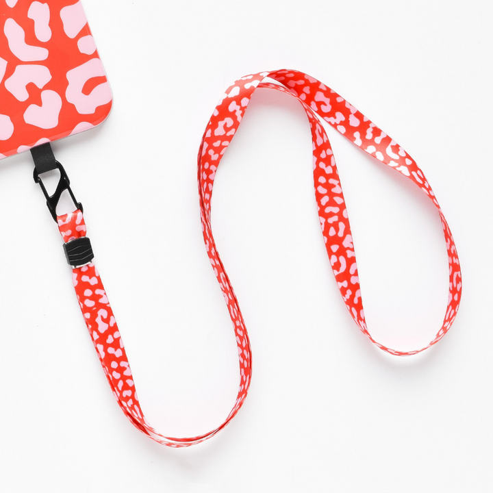 A red Coconut Lane lanyard with a white leopard print pattern is attached to a matching Universal Phone Lanyard Patch Bundle in red and white. The lanyard features a black clasp connecting it to the phone security accessory. The background is a plain white surface.