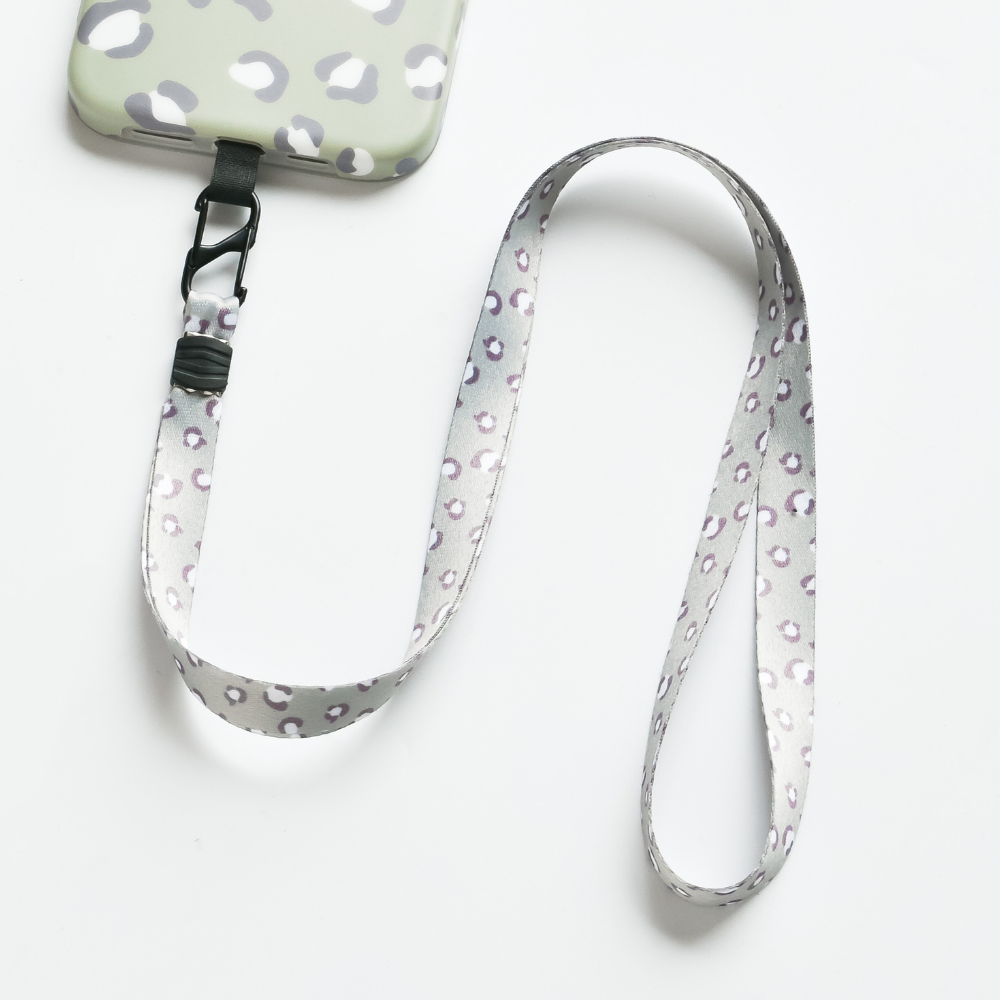 The Coconut Lane Universal Phone Lanyard Patch Bundle features a gray and white phone case adorned with a leopard print pattern, seamlessly attached to a universal phone lanyard patch. The extended lanyard, with a matching gray background and light purple and white leopard print, includes a black clip to securely connect to the case for enhanced phone security.