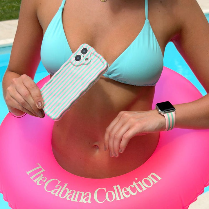 Person in blue bikini holding a matching phone case and watch strap with a blue & pink striped design in a pink inflatable ring labeled 'The Cabana Collection'.
