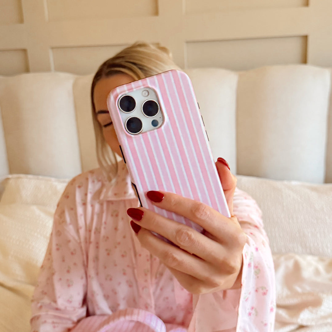 Girl holding a powder stripes phone with a pink case on a bed and pink pjs.