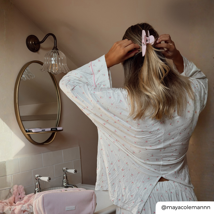 A woman with blonde-highlighted hair seen from behind, wearing a white long-sleeve pajama top with a tiny pink floral pattern and matching shorts. She is adjusting her hair with a light pink claw clip with a small rose detail on it, in a bathroom setting with a sink, mirror.