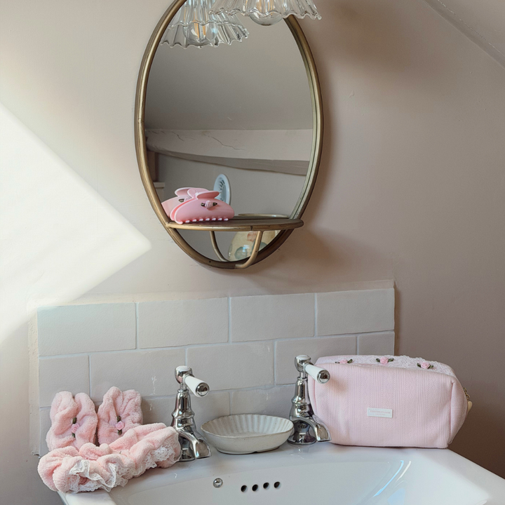 A pink zippered cosmetic pouch with a floral patterned lining sits on a white bathroom sink next to a chrome faucet, with a matching pink headband and wristbands and a gold-framed mirror in the background.