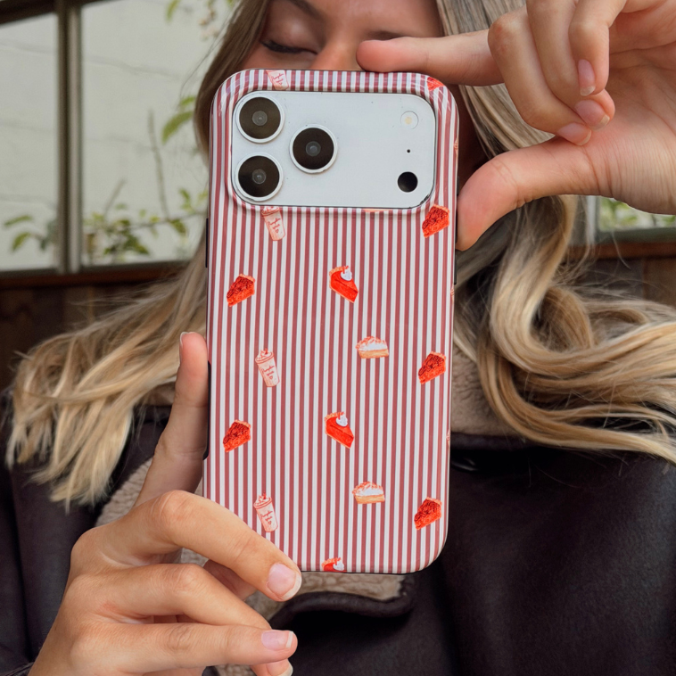 Woman wearing a brown aviator jacket holding up an iphone taking a selfie. The phone is in an autumnal case, with brown vertical stripes, and small prints of pumpkin spice lattes and sweet treats.