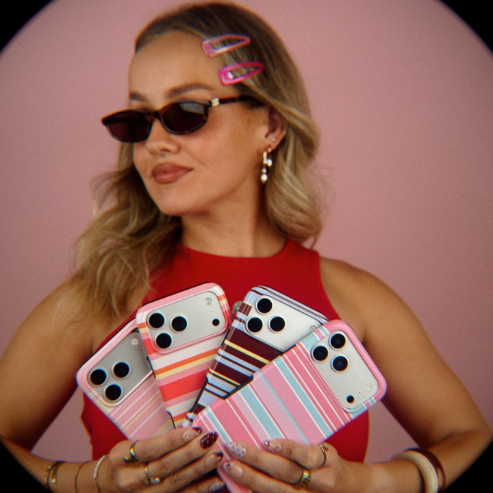 A fish eye effect image of a woman wearing retro sunglasses and old school hair clips holds four striped iPhone cases against a pink background