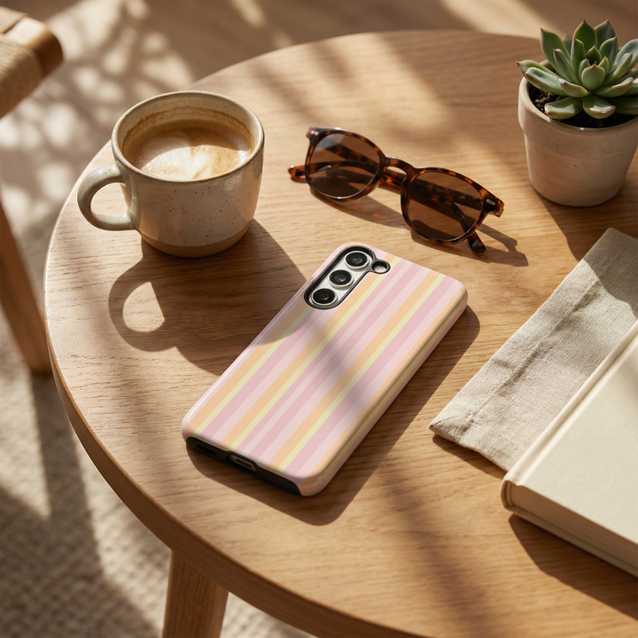Phone case with pastel stripes on a wooden table with a cup of coffee, sunglasses, and a plant.