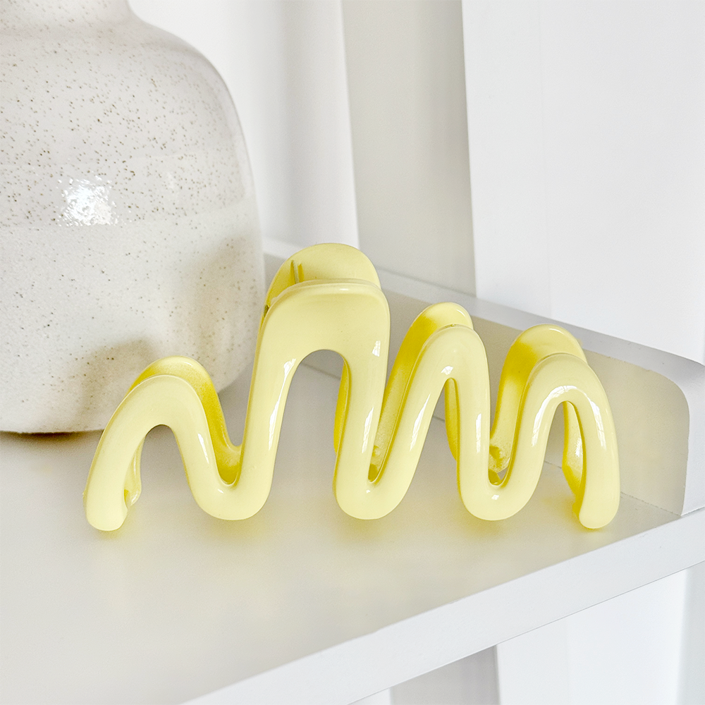 Glossy yellow Coconut Lane Squiggle Claw Clip on a white shelf next to a speckled white vase.
