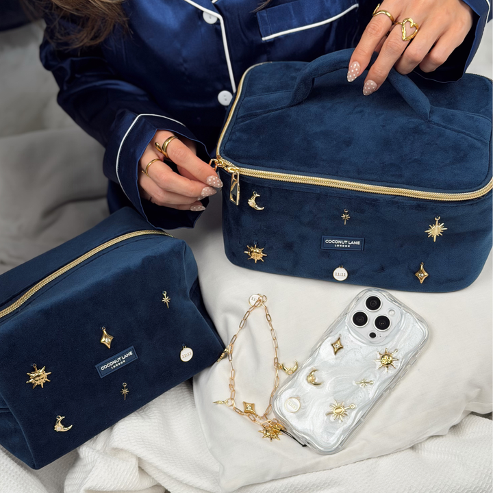 Woman wearing satin navy blue pyjamas. She is unzipping a stardust After Dark Vanity Case. A dark blue navy velvet vanity case with gold zip, and sewn-on gold celestial-themed charms. Including a moon, sun, star, diamond shape and 11:11 charm. Beside the vanity case is the matching makeup bag, phone case and phone strap. 