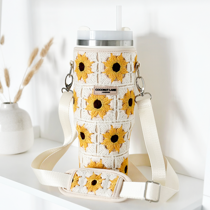 A Coconut Lane Tumbler Carry Case - Sunflower Crochet with a beige strap is displayed on a white shelf. The sleeve showcases yellow sunflowers on white backgrounds. In the background, a small vase containing dried wheat stalks adds a charming rustic touch.