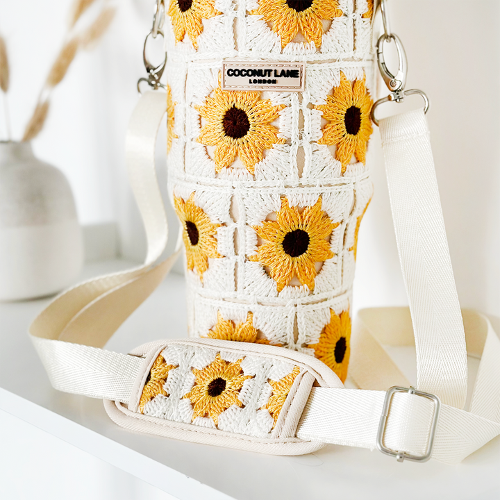 Introducing the Tumbler Carry Case - Sunflower Crochet by Coconut Lane: a charming white crochet crossbody bag embellished with vibrant yellow sunflower patterns. It features a white strap equipped with a padded shoulder section, making it ideal for carrying your Stanley Quencher. This stylish accessory is showcased on a sleek, white surface against a minimalistic, neutral-toned background with a small vase of dried plants nearby.