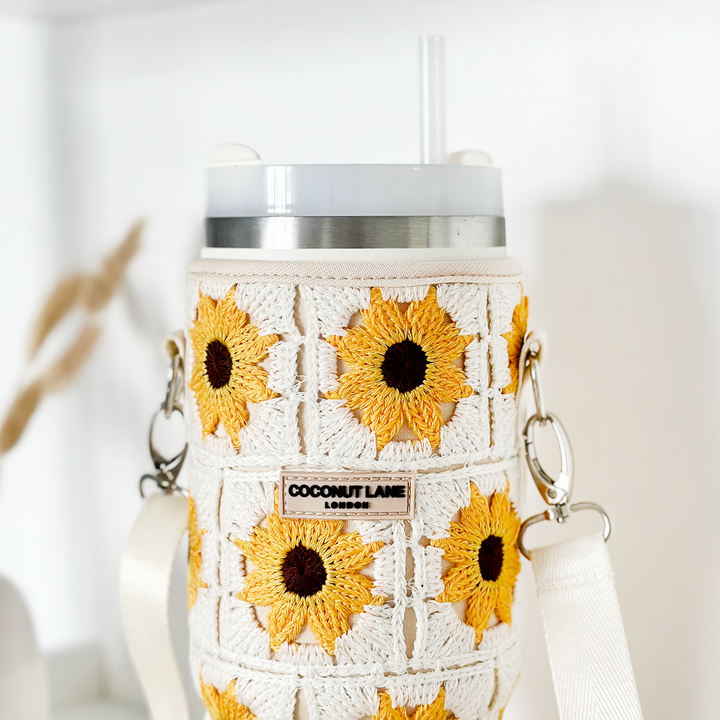A stainless steel Stanley Quencher tumbler with a clear straw is snugly fitted with Coconut Lane's Tumbler Carry Case - Sunflower Crochet. This white crochet sleeve features charming yellow sunflower patterns and a tag that reads "COCONUT LANE." The sleeve also comes with a white strap and silver hooks, making it the perfect travel accessory. The background is white and minimalistic.