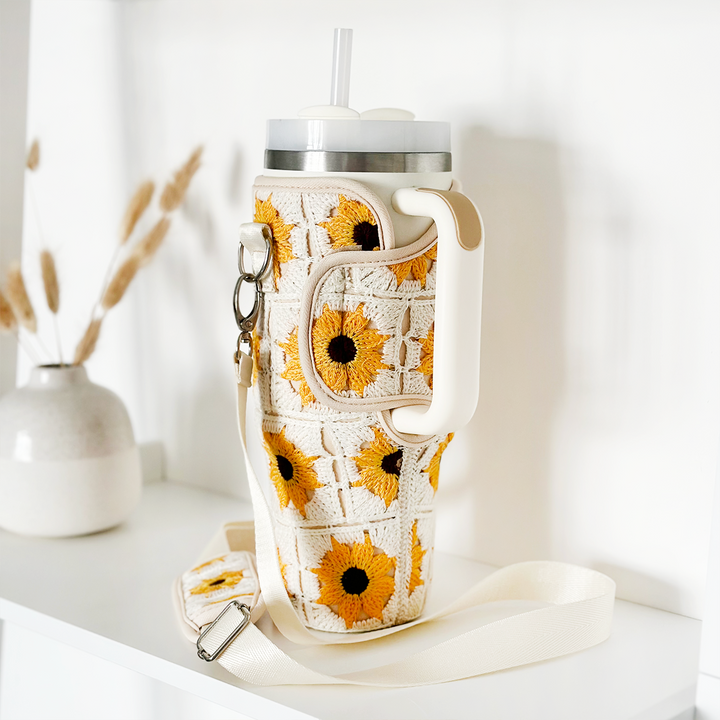 The Coconut Lane Tumbler Carry Case - Sunflower Crochet, a cream-colored crochet water bottle holder adorned with yellow and black sunflower patterns, complete with a white strap, clip, and handle, is displayed on a white shelf with a small vase of dried wheat stalks in the background.