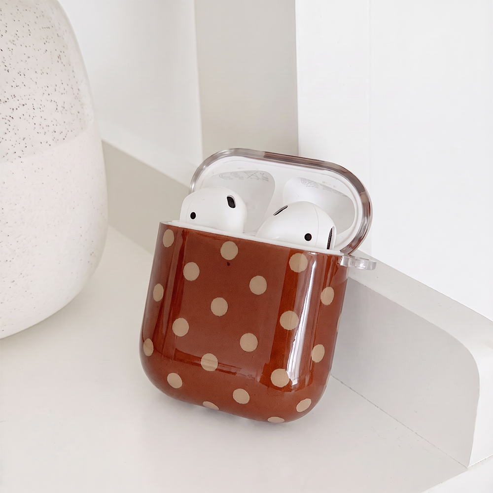 Showing the inside of a glossy brown case for AirPods with cream polka dots and a small clear clip loop on the side. It sits on a white shelf beside a white speckled vase.