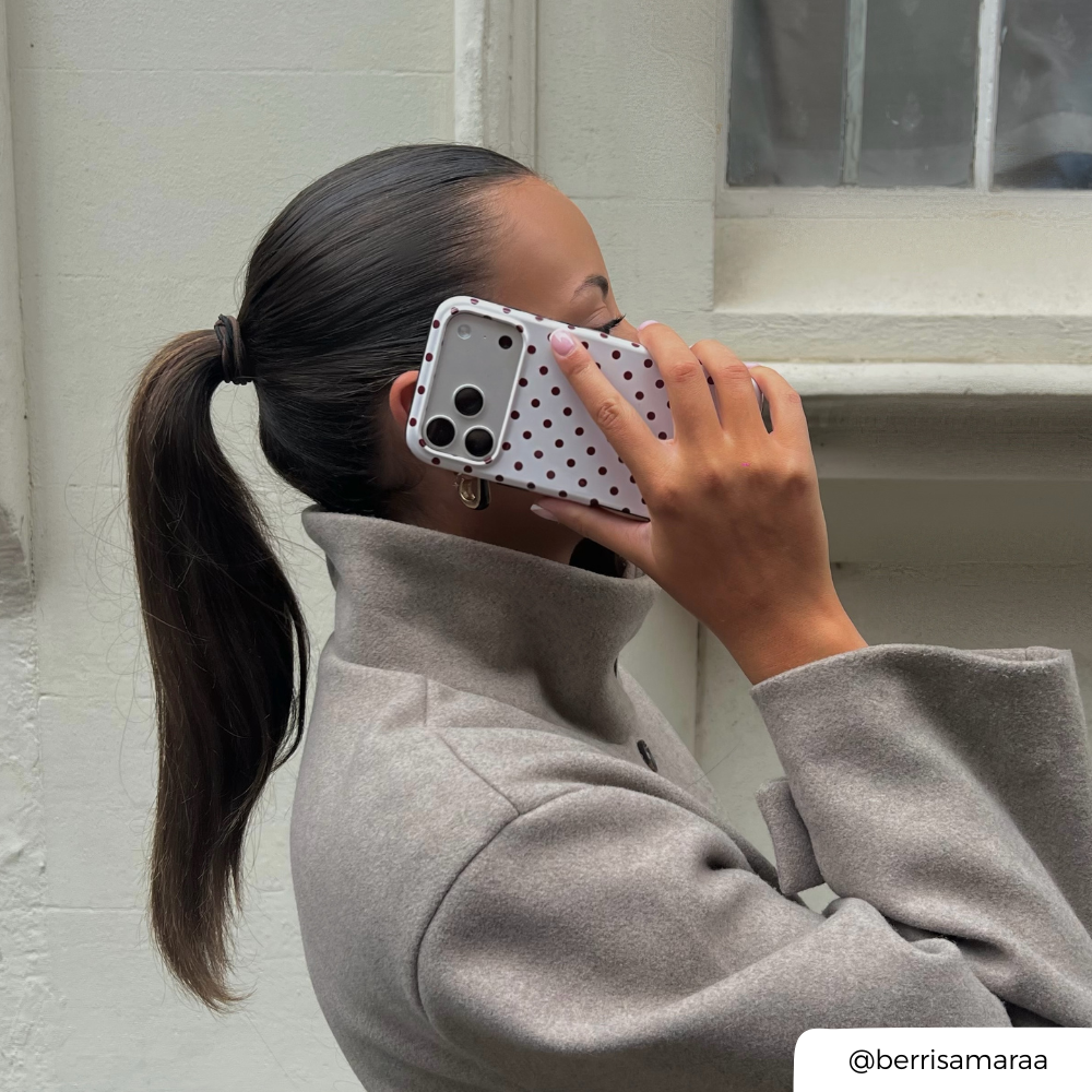Woman with brunette hair tied in a ponytail holds up an iphone to her ear. The phone is in a cream and brown polka dot phone case. 