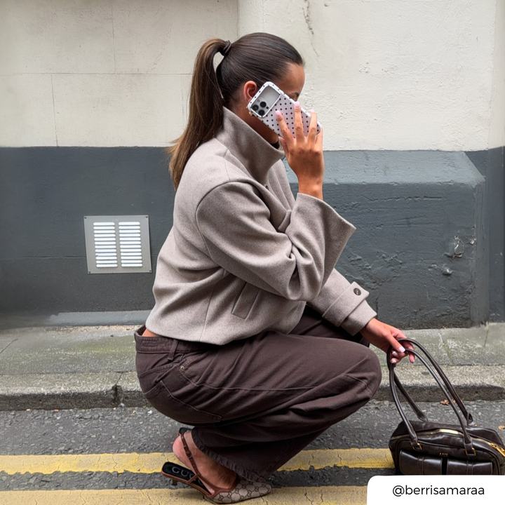 Woman with brunette hair tied in a ponytail holds up an iphone to her ear. The phone is in a cream and brown polka dot phone case. 
