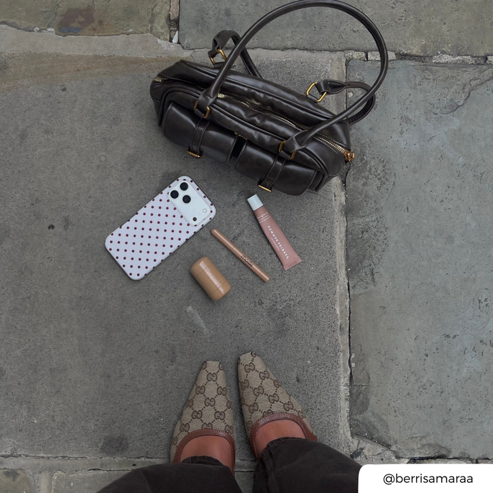 a flat lay photo in the street. A phone in a cream and brown polka dot case placed on the paving slabs next to a brown handbag, and makeup products. 
