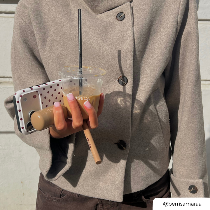 A woman holds an iced coffee, makeup products and phone case in one hand. The phone case is cream with brown polka dots. 