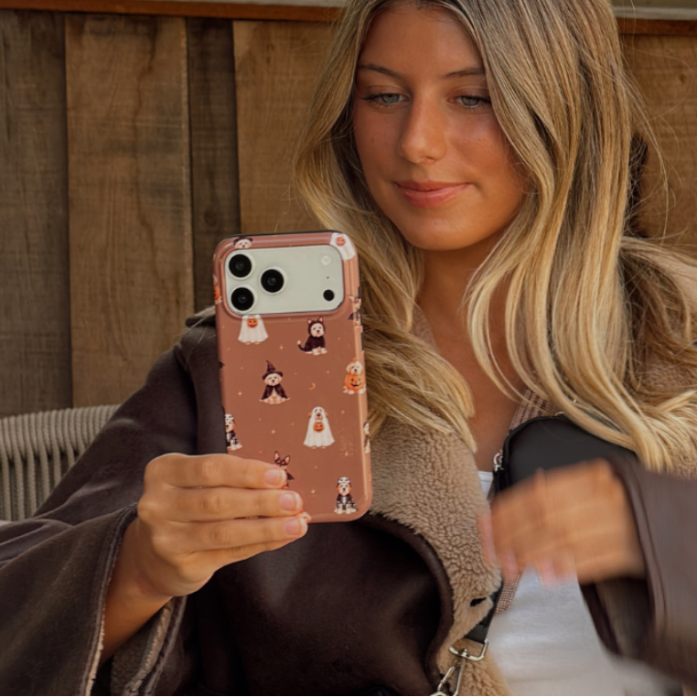 Woman in a brown aviator jacket, holding a phone case with dogs wearing spooky outfits on a brown background.