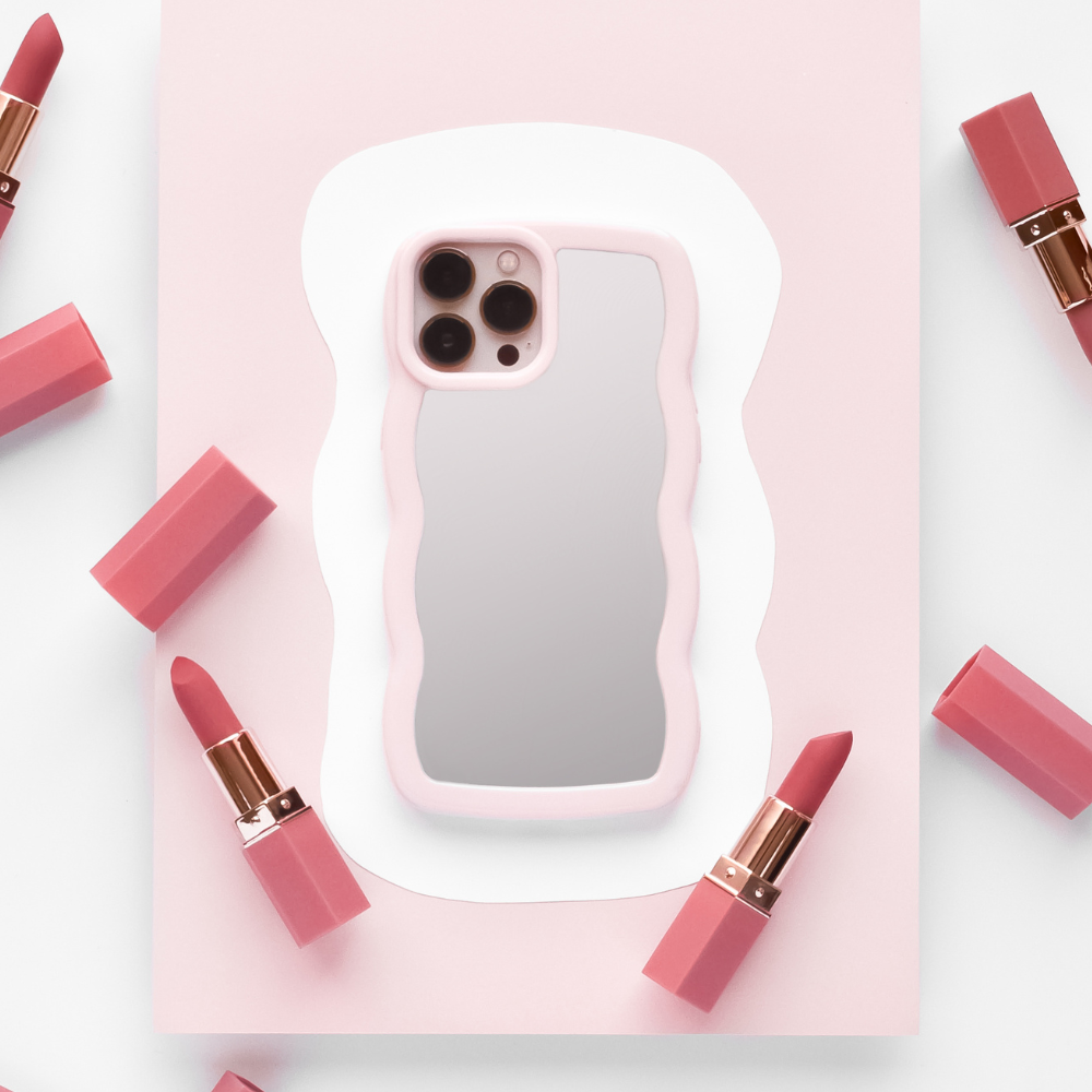 A smartphone featuring the Curvy Mirror Phone Case in Pink by Coconut Lane is centered on a white and pink abstract background. Around the phone, several pink lipsticks, each with a rose gold base and cap, are symmetrically arranged.