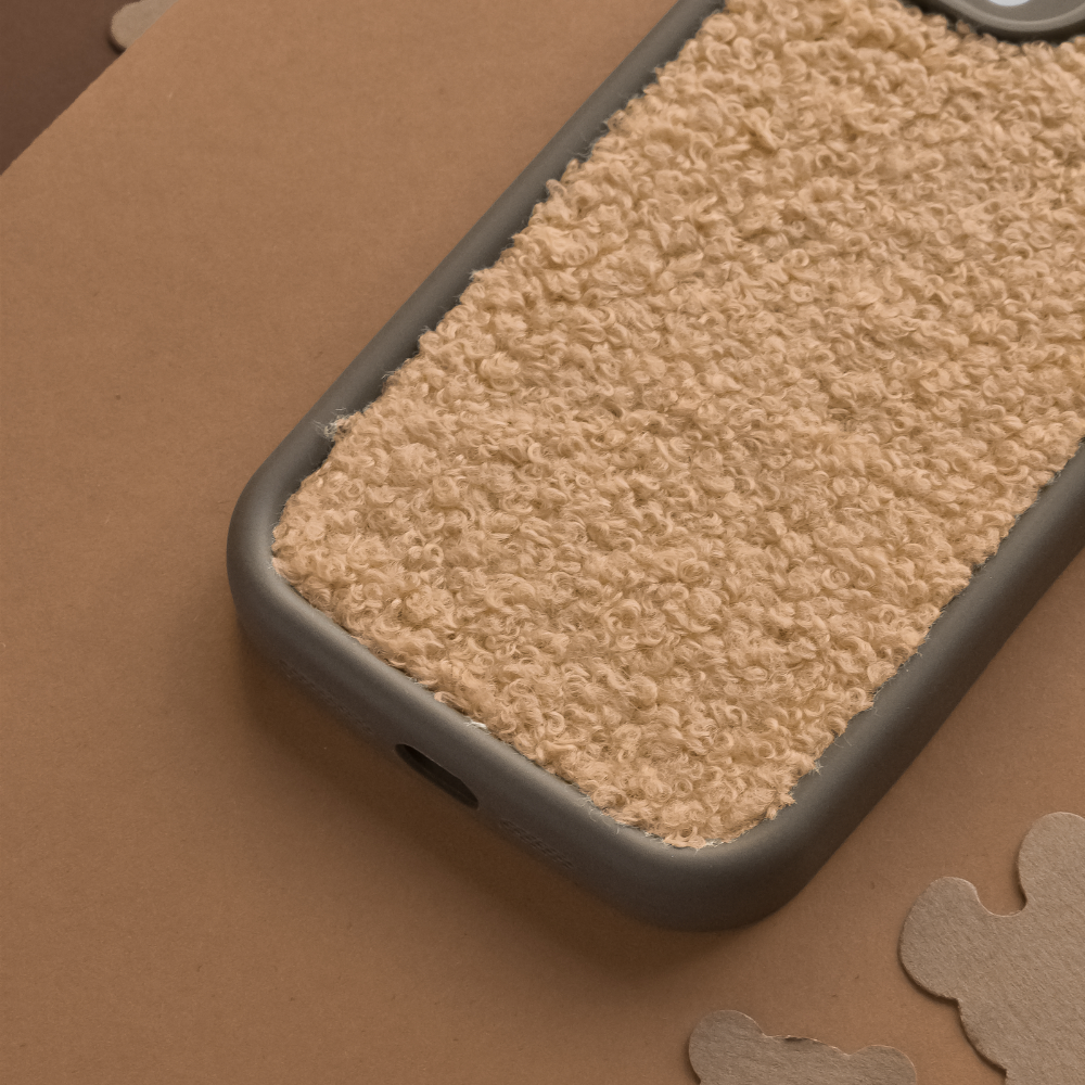 A smartphone with a Cosy Teddy Phone Case - Salted Caramel by Coconut Lane is placed on a brown surface. The case features an extra protective, beige, textured teddy material with a rough, fuzzy appearance and a dark gray border. Several abstract cardboard cutouts are scattered around it.