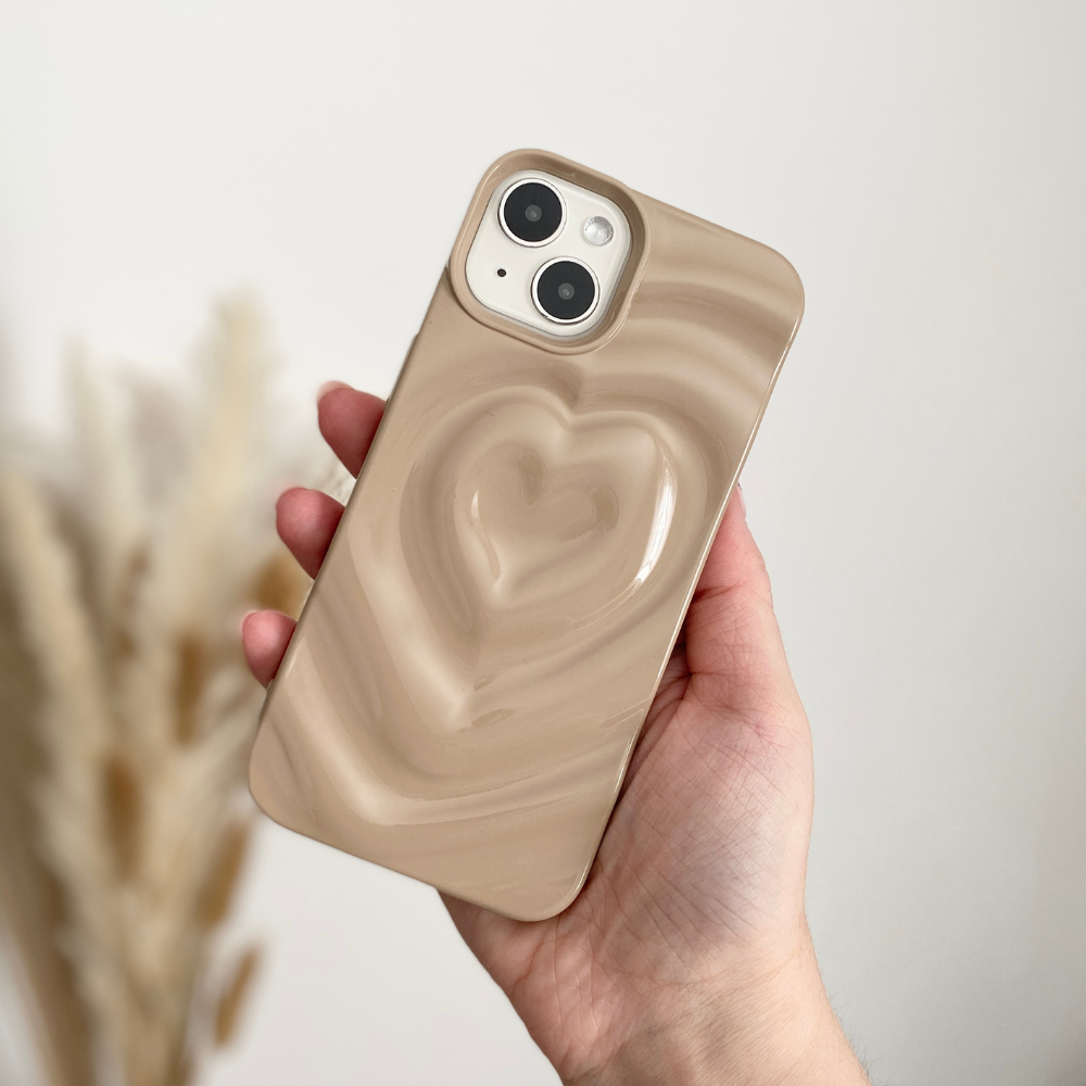 A hand holding an iPhone adorned with Coconut Lane's Melting Heart Phone Case in Biscuit Beige, which features a striking 3D heart design on the back. The background, slightly blurred, highlights some dried pampas grass. This extra-protective phone case effortlessly blends style and durability.