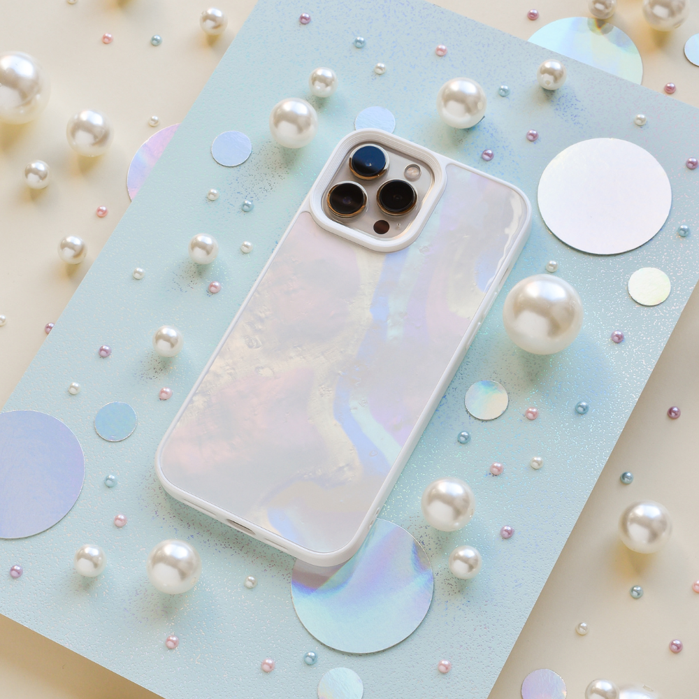An iridescent iPhone with a Coconut Lane Mother of Pearl Phone Case is displayed on a light blue sparkly board. The background is adorned with various sizes of pearls and holographic circles, creating a whimsical and elegant arrangement.