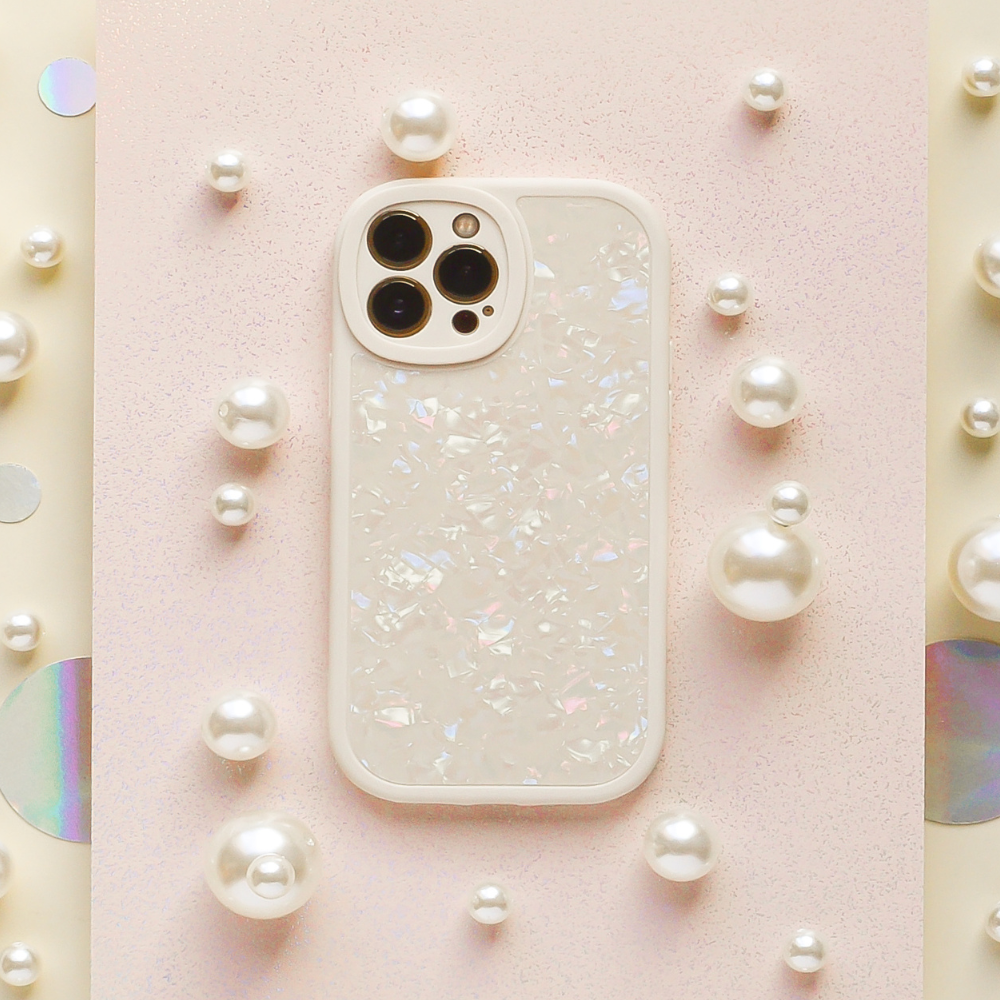 The Moonstone Pearl Phone Case by Coconut Lane adorns a white smartphone with an iridescent shell, which is laid on a pink, textured surface. Surrounding the phone are various-sized pearl-like beads, creating a refined and elegant arrangement. The phone's camera lenses are prominently displayed in the top left corner.
