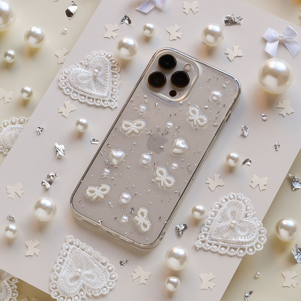 The Coquette Dreams Phone Case by Coconut Lane, crafted as an ultra-protective iPhone 15 case, features a clear base adorned with white lace patterns, pearls, and glitter. It is displayed on a decorated surface that includes scattered white lace hearts, tiny pearls, metallic star-shaped confetti, and small white bows.