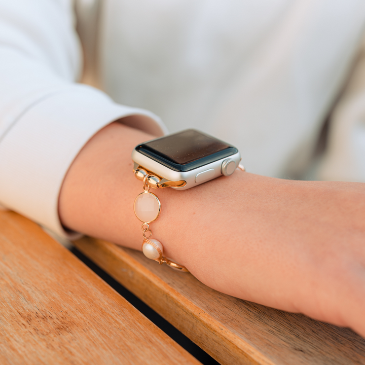 A close-up of a person's wrist showcases the stylish Cute Stone Bracelet Apple Watch Strap by Coconut Lane, featuring a gold chain link band. The person is resting their arm on a wooden surface, adorned with the fashionable strap and wearing a light-colored long sleeve shirt, creating a relaxed and casual appearance.