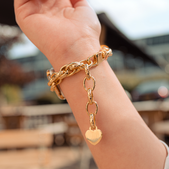 Close-up of a wrist wearing the Coconut Lane Chain Bracelet Apple Watch Strap - Gold, featuring a heart-shaped charm hanging from it. The background is slightly blurred, revealing an outdoor setting with tables and buildings, suggesting a sunny day.