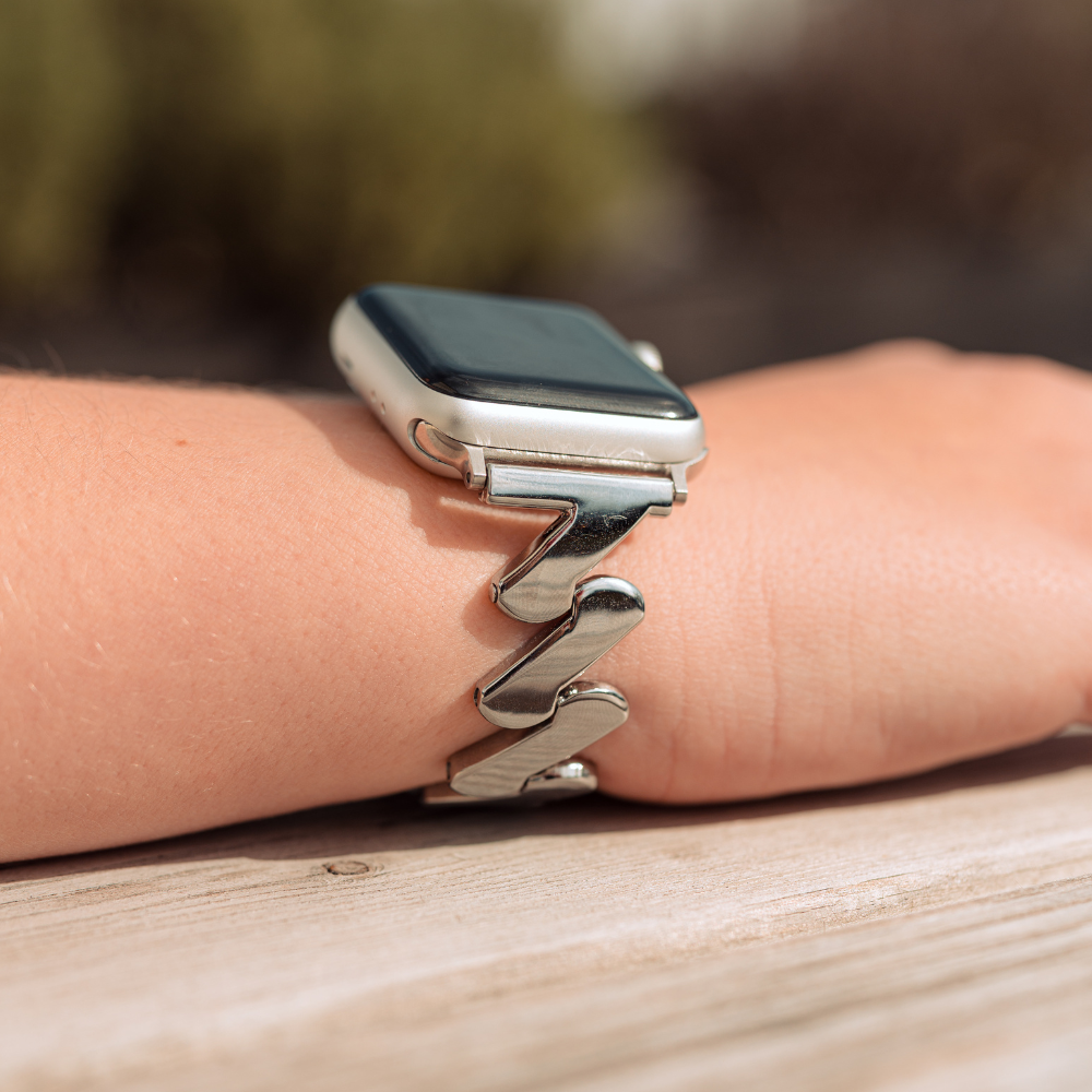A close-up of a silver smartwatch on a person's wrist, featuring Coconut Lane's unique Get Wavy Apple Watch Strap in Silver. The stainless steel band contrasts elegantly with the wooden surface it's resting on, while the background remains slightly blurred.