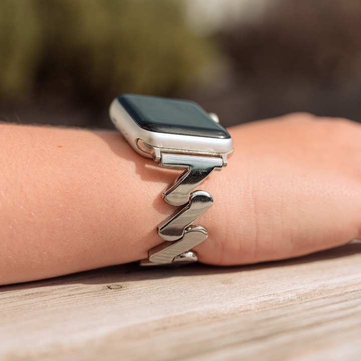 A close-up of a silver smartwatch on a person's wrist, featuring Coconut Lane's unique Get Wavy Apple Watch Strap in Silver. The stainless steel band contrasts elegantly with the wooden surface it's resting on, while the background remains slightly blurred.