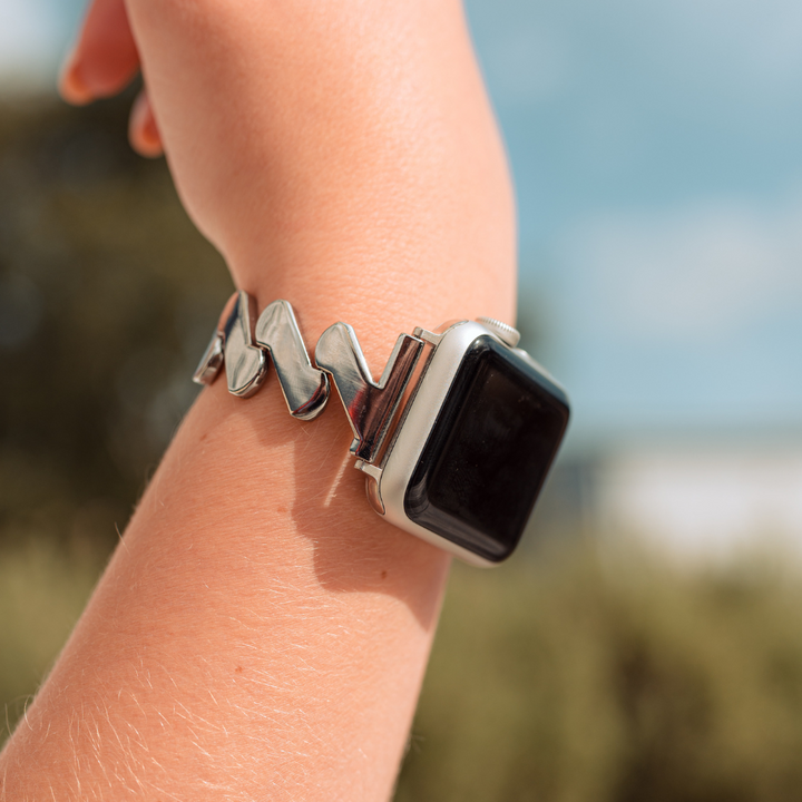 A close-up view of a person wearing a Coconut Lane Get Wavy Apple Watch Strap - Silver, featuring a unique, artistic stainless steel bracelet strap. The silver bracelet consists of interlocking silver-colored shapes. The background is blurred, showcasing natural scenery with greenery and a blue sky.