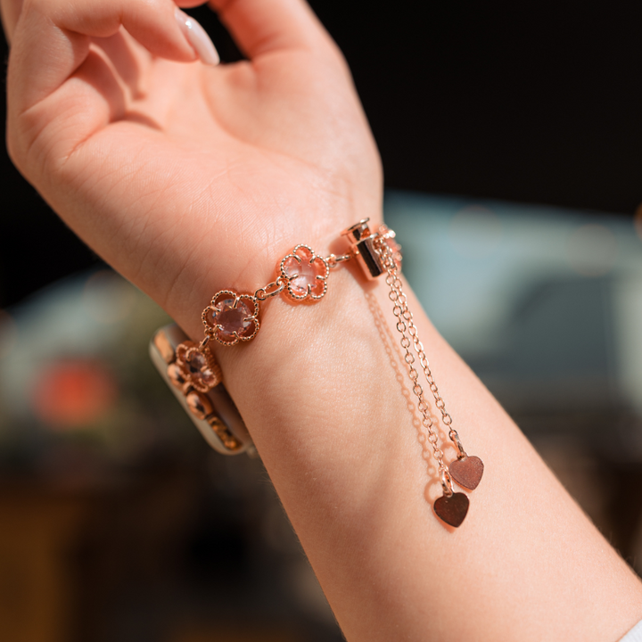 Close-up of a person's wrist adorned with Coconut Lane's Rose Gold Flower Bracelet Apple Watch Strap. The bracelet-like strap highlights several flower-shaped charms and two heart-shaped pendants delicately dangling from the chain. A blurred background enhances the intricate design of this elegant accessory, seamlessly blending style and technology.