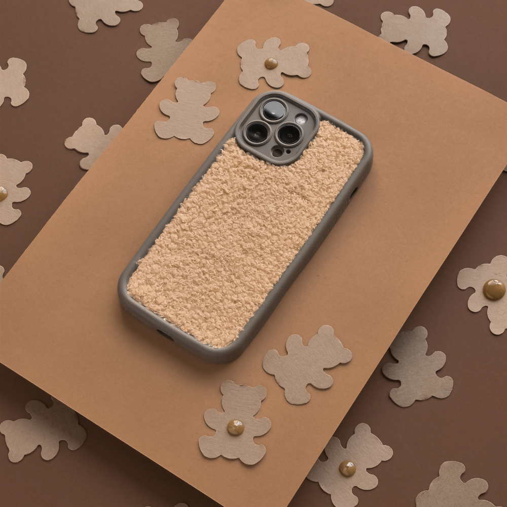 A Cosy Teddy Phone Case - Salted Caramel by Coconut Lane is placed on a brown background. The background is decorated with cut-out shapes of brown teddy bears, adding a playful and cozy vibe to the image.