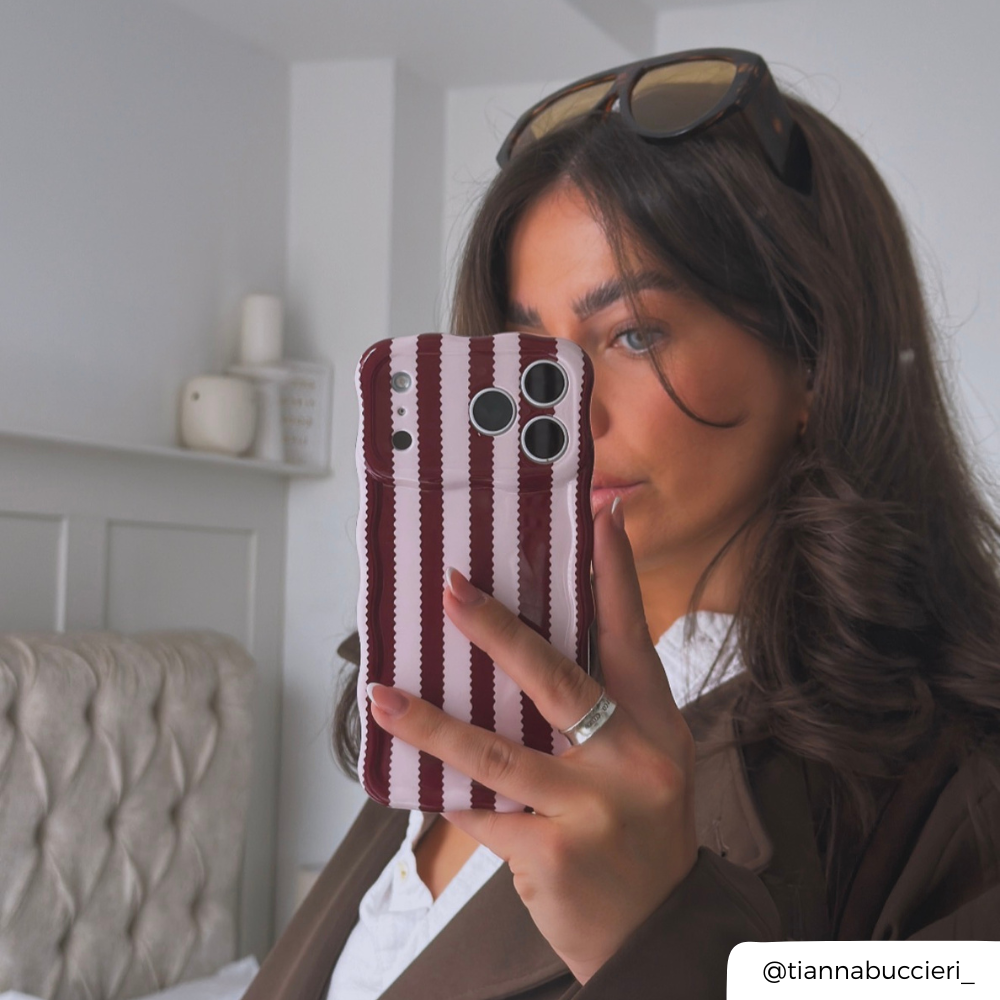 Woman with brunette hair holds up iphone taking a mirror selfie. The phone is in a burgundy and light pink striped wavy phone case.