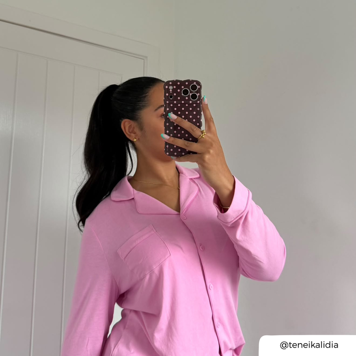 A woman takes a mirror selfie wearing a light pink long-sleeve button-up pyjamas. She is holding a dark brown polka-dot phone case in front of her face with baby pink spots. 